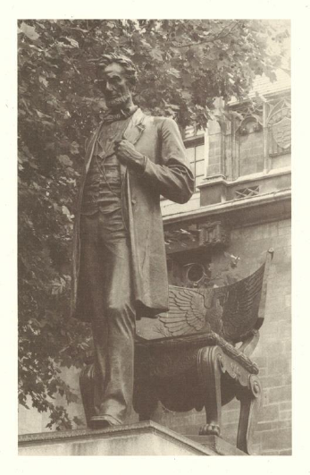 A man in a suit stands outdoors with a book and papers.