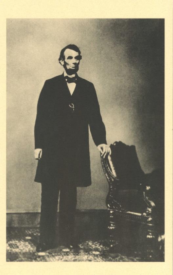 Historical black and white photo of Abraham Lincoln standing beside a chair.